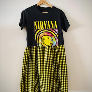 Puede incluir: Vestido camiseta negro con la palabra "NIRVANA" en amarillo y un gráfico de carita sonriente colorido. El vestido tiene una falda a cuadros amarilla y negra, colgado de una percha de madera.