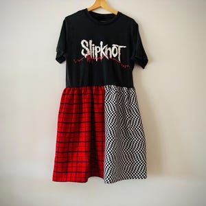 Può includere: Abito nero con logo di una band in alto e gonna con motivo scozzese rosso da un lato e motivo a scacchiera bianco e nero dall'altro. L'abito è appeso a una gruccia di legno.