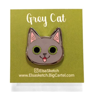 May include: Grey Cat enamel pin with pink ears, green eyes, and a pink tongue sticking out. The pin is on a green background with the text "Grey Cat" and the social media handle "ElsaSketch" and website "www.Elsasketch.BigCartel.com".