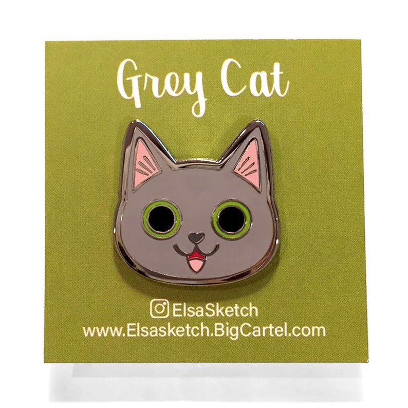 May include: Grey Cat enamel pin with pink ears, green eyes, and a pink tongue sticking out. The pin is on a green background with the text "Grey Cat" and the social media handle "ElsaSketch" and website "www.Elsasketch.BigCartel.com".