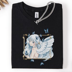 May include: Black t-shirt featuring a framed illustration of a woman with white hair, angel wings, and blue butterflies. A silver chain necklace with a toggle clasp is draped across the shirt.