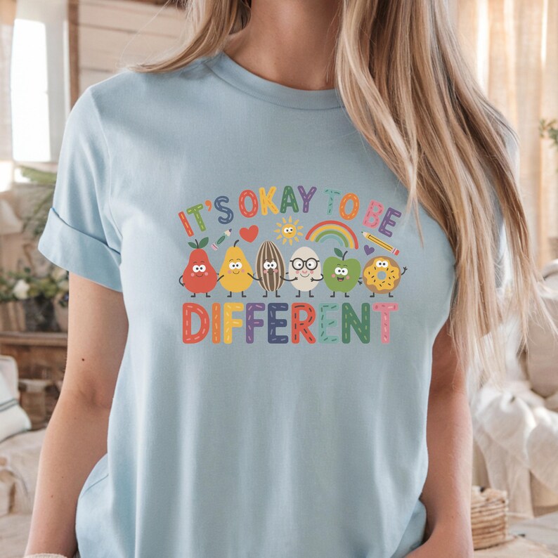 Op de afbeelding: Lichtblauw T-shirt met de tekst "IT'S OKAY TO BE DIFFERENT" in kleurrijke letters. Het shirt heeft een afbeelding van antropomorfe groenten en fruit, waaronder een peer, een hart, een zaad, een ei, een appel en een donut.