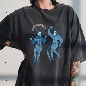 May include: Dark grey t-shirt featuring a whimsical illustration of two blue harlequins with smiling faces and star patterns. One harlequin wears a hat, the other has horns. A rainbow arcs above them. The design is printed on a soft, vintage-style tee.