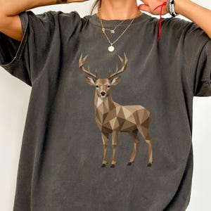 Może przedstawiać: Ciemnoszary t-shirt z krótkim rękawem z geometryczną ilustracją w stylu low-poly przedstawiającą brązowego jelenia z dużym porożem, umieszczoną na środku. Koszulka ma okrągły dekolt i luźny krój w stonowanej, jednolitej kolorystyce.