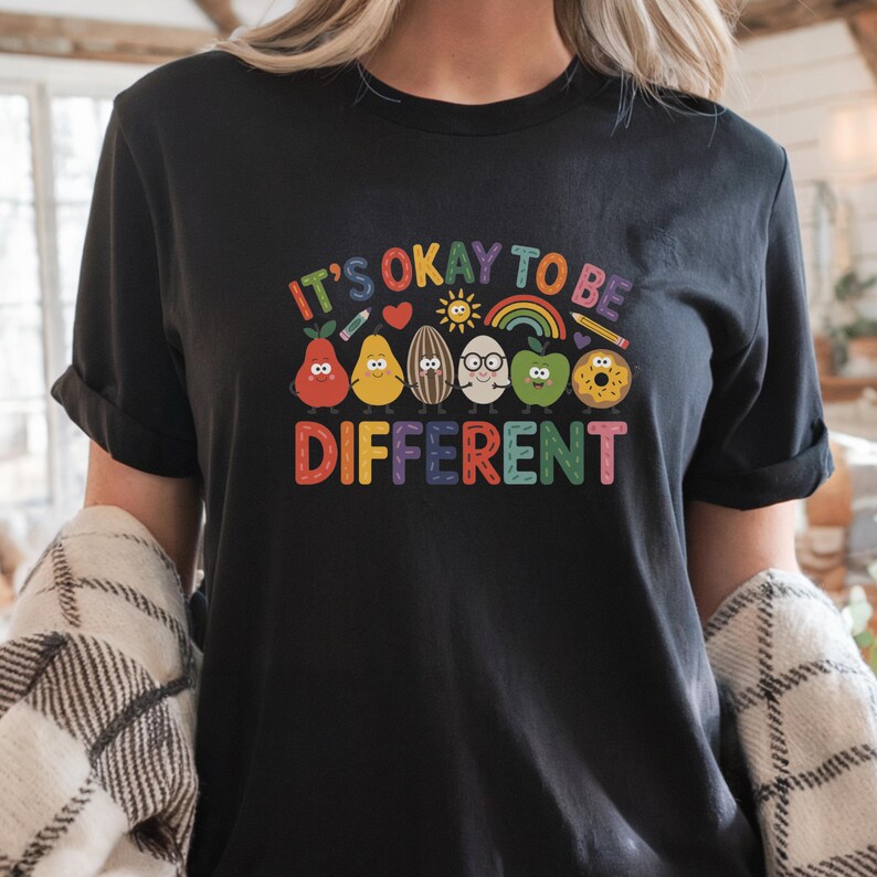 Op de afbeelding: Zwart t-shirt met de kleurrijke tekst "IT'S OKAY TO BE DIFFERENT". De afbeelding toont cartoon fruit en groenten met lachende gezichten, een regenboog en een zon.