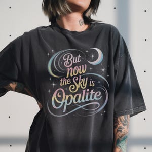 Könnte beinhalten: Dunkelgraues T-Shirt mit dem Text "But now the Sky is Opalite" in einem farbenfrohen, himmlischen Design. Das Design umfasst einen Halbmond, Sterne und eine Regenbogen-Farbpalette. Das Shirt hat einen Vintage-Look.
