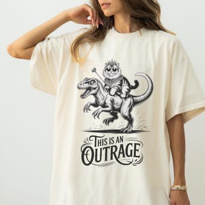 May include: Cream-colored t-shirt featuring a black and white illustration of a cat wearing a crown and riding a dinosaur. The text "THIS IS AN OUTRAGE" is below the illustration. The shirt has short sleeves and a relaxed fit.