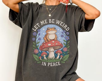 May include: Dark gray t-shirt with the text "LET ME BE WEIRD IN PEACE." The shirt features a whimsical illustration of a frog sitting on a red and white mushroom, surrounded by flowers and foliage.