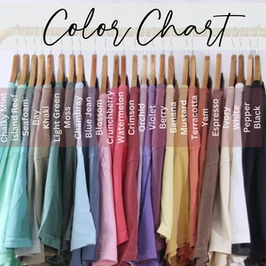 Op de afbeelding: Een kleurenkaart toont een verscheidenheid aan t-shirts in verschillende kleuren, waaronder Chalky Mint, Island Reef, Seafoam, Bay, Khaki en meer. De shirts hangen aan houten hangers op een gouden rek. De tekst "Color Chart" staat bovenaan.