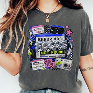 May include: A charcoal grey t-shirt featuring a graphic print of computer error windows, headphones, and a digital camera. The text reads ERROR 404: FOCUS NOT FOUND, DISTRACTED, ATTENTION DISCONNECTED, and PLEASE WAIT. The design includes pixelated stars.