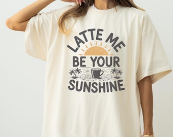 以下が含まれることがあります： 「LATTE ME BE YOUR SUNSHINE」の文字が黒とオレンジでプリントされた、クリーム色のTシャツ。デザインには太陽、ヤシの木、貝殻、コーヒーカップが含まれています。カジュアルでリラックスしたフィット感。