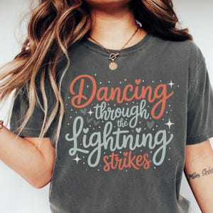 May include: Dark grey t-shirt featuring the text "Dancing through the Lightning strikes" in coral and light blue script. The design includes hearts and stars. The shirt has a crew neck and short sleeves.