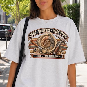 Könnte beinhalten: Ein hellgraues T-Shirt mit einem Grafikdruck eines Ammoniten-Fossils, eines Pinsels und eines Geologenhammers. Der Text lautet DUST BRUSHES DEEP TIME und FOSSIL PREP FIELD CREW. Das Design zeigt geschichtete Gesteinsschichten in Rot, Gelb und Blau.