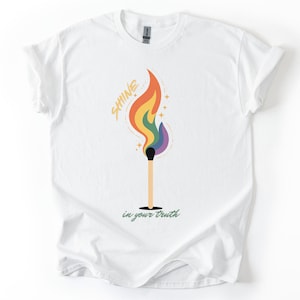 May include: White t-shirt featuring a colorful graphic of a lit match with a rainbow flame. The text "SHINE" is above the flame, and "in your truth" is below. The design includes yellow stars.