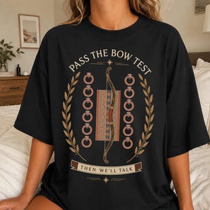 May include: A black t-shirt featuring a graphic print with the text "PASS THE BOW TEST" at the top and "THEN WE'LL TALK" at the bottom. The design includes a central bow, a patterned rectangular block, two columns of rings, and laurel leaf branches.