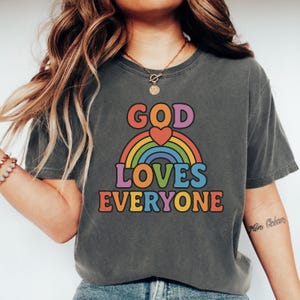 May include: A charcoal grey t-shirt with a rainbow and the text "God Loves Everyone" in colorful letters. A red heart is in the center of the rainbow.