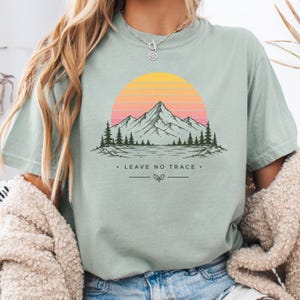 May include: A sage green t-shirt featuring a graphic of a mountain range and pine trees set against a sunset with horizontal orange and pink stripes. Below the landscape, the text reads LEAVE NO TRACE with a small leaf icon and horizontal lines.