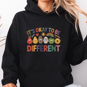 Op de afbeelding: Zwarte hoodie met de tekst "IT'S OKAY TO BE DIFFERENT" in kleurrijke letters. Het ontwerp toont cartoon fruit en groenten met gezichten, waaronder een peer, citroen, avocado, ei, appel en donut. Een regenboog en een zon zijn ook inbegrepen.