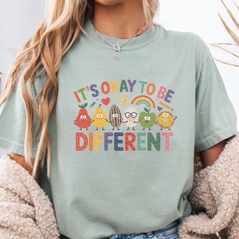 Op de afbeelding: Lichtgroen T-shirt met de tekst "IT'S OKAY TO BE DIFFERENT" in kleurrijke letters. Het shirt heeft een afbeelding van lachende fruitkarakters, waaronder een peer, een citroen, een zonnebloempit, een bril, een appel en een donut.