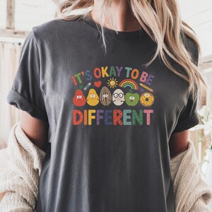 Op de afbeelding: Donkergrijs T-shirt met de kleurrijke tekst "IT'S OKAY TO BE DIFFERENT". Het shirt heeft cartoon illustraties van verschillende voedingsmiddelen met gezichten, waaronder een peer, een citroen, een ei en een appel.