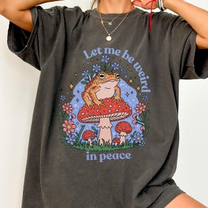 Può includere: Una maglietta grigia scura con una grafica di una rana seduta su un fungo. Il testo "Let me be weird in peace" è stampato sulla maglietta.