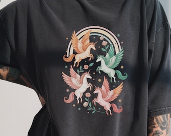 May include: Dark gray t-shirt featuring a colorful illustration of four winged unicorns. The unicorns have various colors, including peach, white, and green, with a rainbow and floral accents. The design is centered on the shirt.