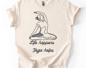 May include: Cream-colored t-shirt featuring a line drawing of a person in a yoga pose. The text "Life happens Yoga helps" is printed in black script below the illustration. The shirt has short sleeves and a crew neck.