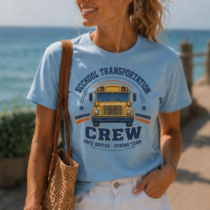 May include: A light blue short-sleeve t-shirt featuring a graphic of a yellow school bus. The text reads "SCHOOL TRANSPORTATION CREW" in dark blue, with "SAFE ROUTES" and "STRONG TEAM" printed below the bus in a circular design.