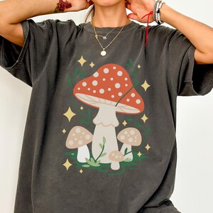 Op de afbeelding: Donkergrijs T-shirt met een paddestoelontwerp. De afbeelding toont een grote rode paddestoel met witte stippen, kleinere beige paddestoelen, groene bladeren en gele sterren. Het T-shirt heeft een relaxte pasvorm.