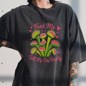 May include: Dark gray t-shirt featuring a graphic of Venus flytraps in green and pink, with the text "Feed Me" and "Tell Me I'm Pretty" in pink. The design includes pink hearts.