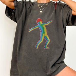 Puede incluir: Camiseta gris oscuro con una ilustración colorida de una persona con los brazos extendidos. La figura está representada en un degradado de colores arcoíris, con un gorro rojo. La camiseta tiene un corte holgado y un aspecto vintage.