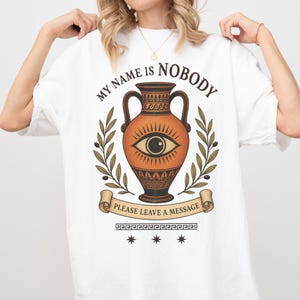 Könnte beinhalten: Ein weißes T-Shirt mit einem Grafikdruck einer orange-schwarzen Amphore im griechischen Stil mit einem Auge in der Mitte. Der Text MY NAME IS NOBODY steht über der Vase und PLEASE LEAVE A MESSAGE auf einem Banner darunter, flankiert von Olivenzweigen.