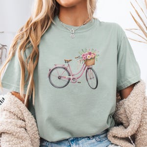 Puede incluir: Camiseta verde claro con una ilustración en acuarela de una bicicleta rosa. La bicicleta tiene una cesta llena de flores rosas y moradas y un asiento marrón. La camiseta tiene mangas cortas.