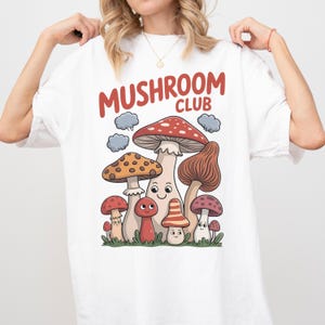 May include: White t-shirt with a colorful mushroom graphic. The design features various cartoon mushrooms with the text "MUSHROOM CLUB" in red. The mushrooms are in shades of red, brown, and orange, with green grass at the bottom.