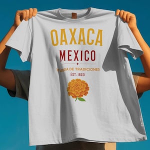 May include: A light gray short-sleeve t-shirt featuring the text OAXACA MEXICO in yellow and dark red, with the phrase TIERRA DE TRADICIONES EST. 1823 below it. A graphic of an orange marigold flower with green leaves is centered at the bottom.