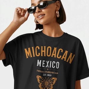 Op de afbeelding: Een zwart t-shirt met de tekst MICHOACAN in grote oranje letters, MEXICO in wit, en TIERRA PUREPECHA EST. 1823 in kleinere oranje tekst. Onder de tekst staat een afbeelding van een oranje vlinder met zwarte omlijningen en details.