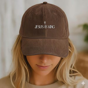Puede incluir: Gorra de béisbol marrón con el texto "JESUS IS KING" en blanco. La gorra presenta una cruz sobre el texto y tiene una visera curva. La gorra está hecha de una tela texturizada.