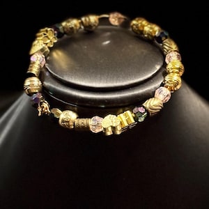 Puede incluir: Una pulsera de cuentas doradas con una variedad de formas y tamaños de cuentas. La pulsera presenta cuentas redondas, cilíndricas y en forma de flor, intercaladas con cuentas de cristal facetadas en tonos rosas y morados. La pulsera se muestra sobre un fondo oscuro.