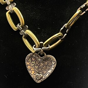 K&ouml;nnte beinhalten: Eine gold- und silberfarbene Halskette mit Herzanh&auml;nger. Die Kette hat abwechselnd goldene und silberne Glieder. Der Herzanh&auml;nger ist silberfarben und mit kleinen Herzformen bedeckt.