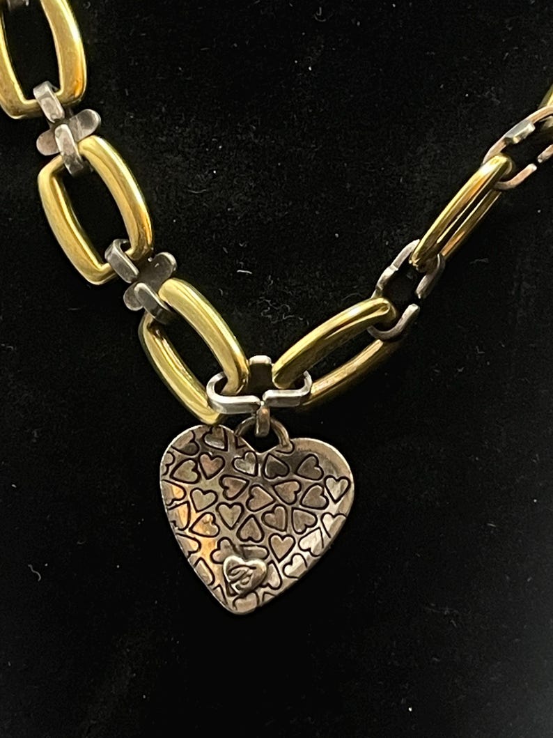 K&ouml;nnte beinhalten: Eine gold- und silberfarbene Halskette mit Herzanh&auml;nger. Die Kette hat abwechselnd goldene und silberne Glieder. Der Herzanh&auml;nger ist silberfarben und mit kleinen Herzformen bedeckt.