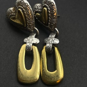 K&ouml;nnte beinhalten: Ein Paar Ohrh&auml;nger mit herzf&ouml;rmigem Oberteil und einem goldfarbenen, offenen ovalen Tropfen. Das herzf&ouml;rmige Oberteil hat ein silberfarbenes, verziertes Design mit einem goldfarbenen Herz in der Mitte. Die Ohrringe sind auf schwarzem Hintergrund.