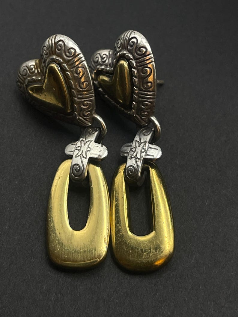 K&ouml;nnte beinhalten: Ein Paar Ohrh&auml;nger mit herzf&ouml;rmigem Oberteil und einem goldfarbenen, offenen ovalen Tropfen. Das herzf&ouml;rmige Oberteil hat ein silberfarbenes, verziertes Design mit einem goldfarbenen Herz in der Mitte. Die Ohrringe sind auf schwarzem Hintergrund.
