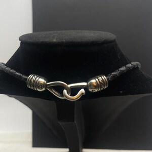 K&ouml;nnte beinhalten: Schwarze Lederkette mit geflochtenem Design. Die Halskette hat silberfarbene Metallakzente und einen Hakenverschluss. Die Halskette wird auf einer schwarzen Schmuckb&uuml;ste vor einem schwarz-wei&szlig;en Hintergrund pr&auml;sentiert.