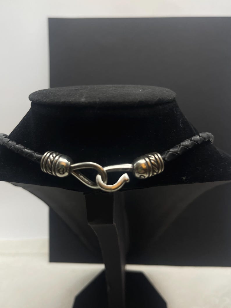 K&ouml;nnte beinhalten: Schwarze Lederkette mit geflochtenem Design. Die Halskette hat silberfarbene Metallakzente und einen Hakenverschluss. Die Halskette wird auf einer schwarzen Schmuckb&uuml;ste vor einem schwarz-wei&szlig;en Hintergrund pr&auml;sentiert.