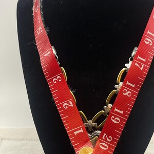 K&ouml;nnte beinhalten: Eine goldfarbene Halskette mit silbernen Akzenten und einem schwarzen Lederband. Die Halskette wird auf einer schwarzen Schaufensterpuppe pr&auml;sentiert. Ein rotes Ma&szlig;band ist &uuml;ber die Halskette gelegt und zeigt ein Ma&szlig; von ca. 53 cm.