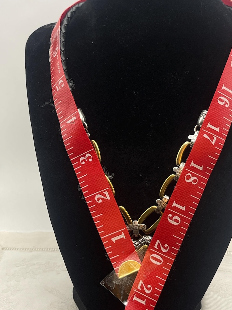 K&ouml;nnte beinhalten: Eine goldfarbene Halskette mit silbernen Akzenten und einem schwarzen Lederband. Die Halskette wird auf einer schwarzen Schaufensterpuppe pr&auml;sentiert. Ein rotes Ma&szlig;band ist &uuml;ber die Halskette gelegt und zeigt ein Ma&szlig; von ca. 53 cm.