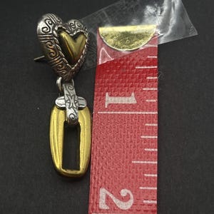 K&ouml;nnte beinhalten: Ein silberner und goldfarbener herzf&ouml;rmiger Ohrring mit einem offenen rechteckigen Anh&auml;nger. Der Ohrring ist etwa 3,8 cm lang. Das Herz hat ein verziertes Design und eine goldfarbene Mitte. Der Anh&auml;nger ist goldfarben mit silbernem Akzent.
