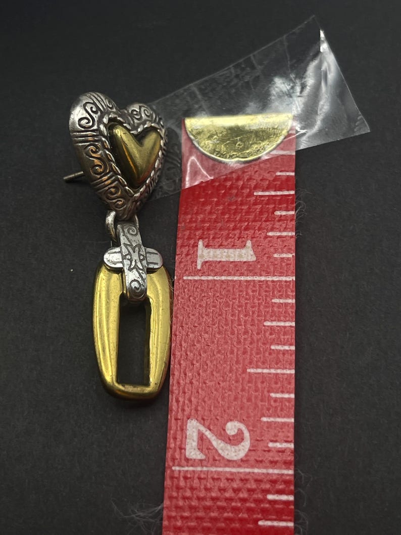 K&ouml;nnte beinhalten: Ein silberner und goldfarbener herzf&ouml;rmiger Ohrring mit einem offenen rechteckigen Anh&auml;nger. Der Ohrring ist etwa 3,8 cm lang. Das Herz hat ein verziertes Design und eine goldfarbene Mitte. Der Anh&auml;nger ist goldfarben mit silbernem Akzent.