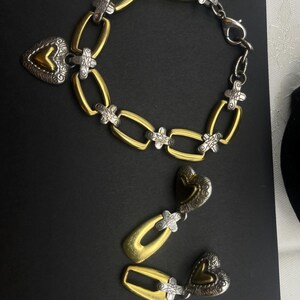 K&ouml;nnte beinhalten: Ein gold- und silberfarbenes Armband und ein passendes Ohrring-Set. Das Armband hat ovale Glieder mit silbernen Akzenten und einen herzf&ouml;rmigen Anh&auml;nger. Die Ohrringe haben ein &auml;hnliches Design mit gold- und silberfarbenen herzf&ouml;rmigen Anh&auml;ngern.