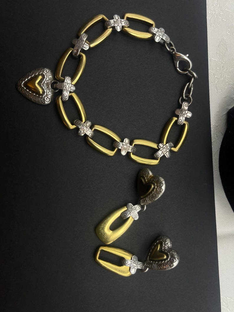 K&ouml;nnte beinhalten: Ein gold- und silberfarbenes Armband und ein passendes Ohrring-Set. Das Armband hat ovale Glieder mit silbernen Akzenten und einen herzf&ouml;rmigen Anh&auml;nger. Die Ohrringe haben ein &auml;hnliches Design mit gold- und silberfarbenen herzf&ouml;rmigen Anh&auml;ngern.